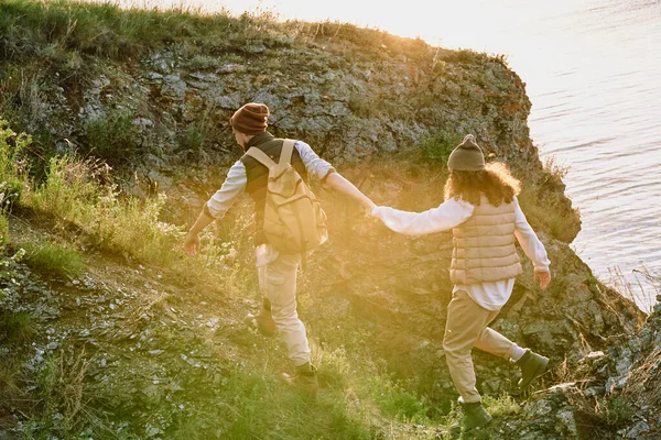 年轻的男性徒步旅行者 背着背包牵着他的妻子 夕阳西下 他们都走在绿草覆盖的岩石上 — 图库照片