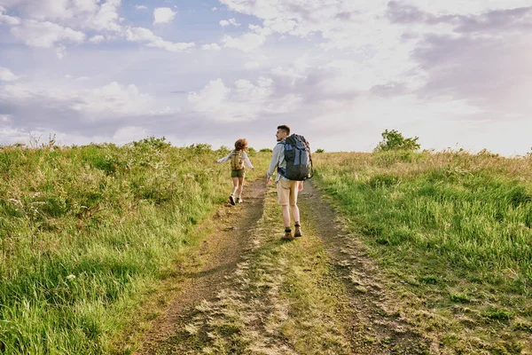 年轻的女徒步旅行者背着背包在乡间路上奔跑 而她的丈夫背着背包站在后面望着旁边的背影 — 图库照片