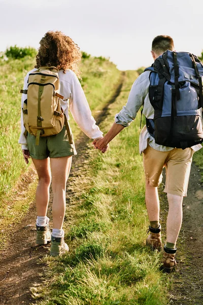 两个年轻多情的徒步旅行者手提着背包 走在绿草覆盖的乡间路上的背景图 — 图库照片