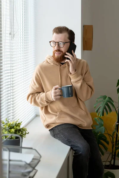 Gülümseyen, kapüşonlu, genç, sakallı adam pencere eşiğinde oturuyor ve cep telefonuyla konuşurken kahve içiyor.