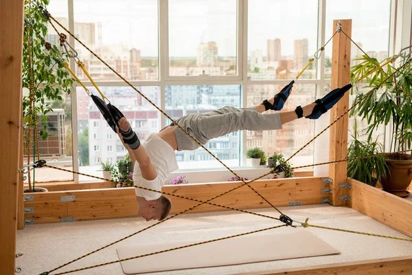 Young contemporary sportsman with his arms and legs fixed to ropes doing difficult aerial yoga exercises while hanging over the floor