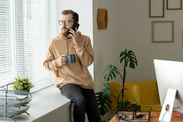 窓ガラスの上に座って 電話で通信しながらブラインドを通して見ている深刻な若い髭の男 — ストック写真