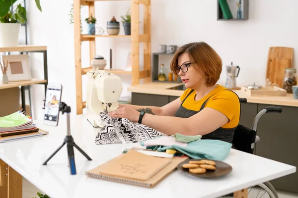 Çevrimiçi izleyicileri için ana kurs sırasında akıllı telefon kamerasının önünde tekerlekli sandalyede oturan hoş genç terzi.