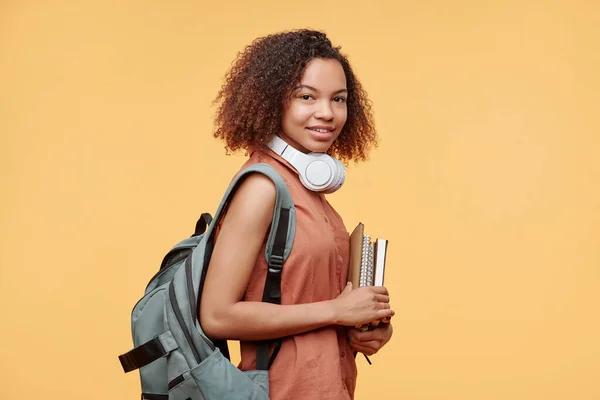 Portrait of smiling Black student girl with Afro hairstyle standing with workbooks against yellow background