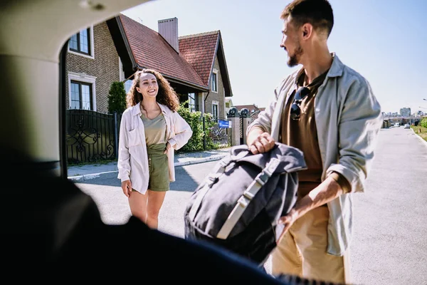 明るい夏の朝を楽しみながら 彼女の夫を見て陽気若い女性は車のトランクに大きなバックパックを配置しようとします — ストック写真