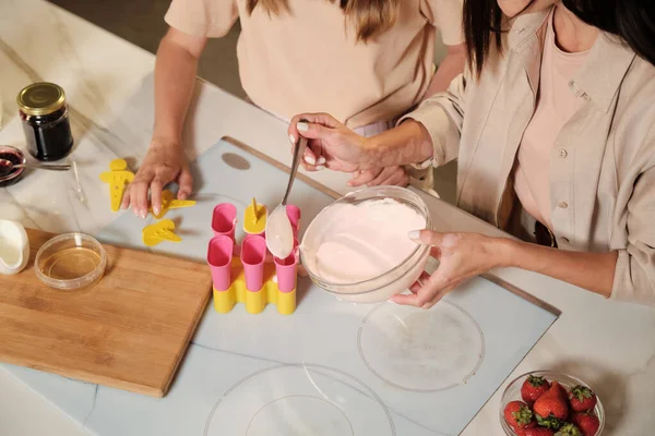 Elinde kaşık olan genç bir kadının elleri ev yapımı dondurmanın karışımını silikon formlara yerleştiriyor ve kızı yanında duruyor.