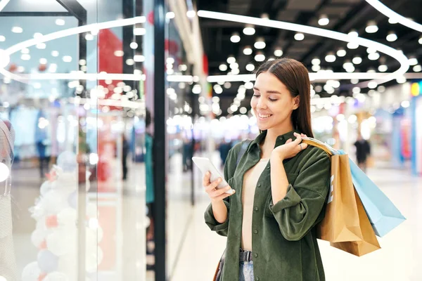 Young cheerful brunette shopper with paperbags scrolling in smartphone or looking through messages or adverts while shopping in the mall