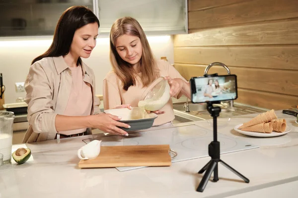 Blond teenage girl pouring mixture of homemade icecream ingredients into large bowl in front of smartphone camera in the kitchen