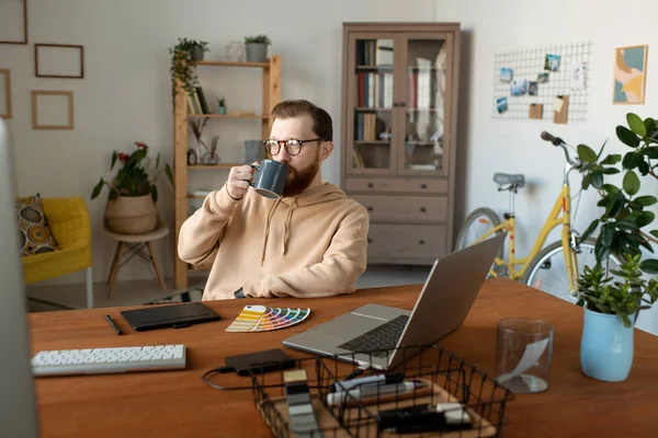コンピュータと自宅のオフィスでコーヒーを飲みながら机に座って穏やかなハンサムなひげそりのデザイナー — ストック写真