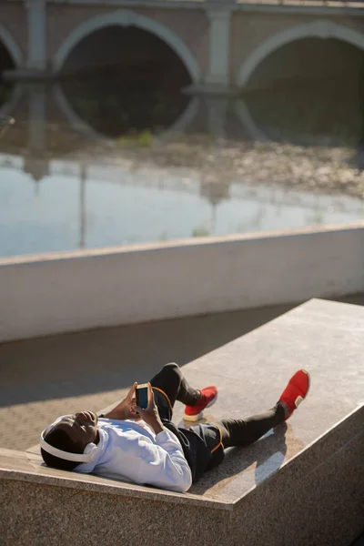 Joven Hombre Africano Relajado Ropa Deportiva Auriculares Blancos Tumbado Orillas — Foto de Stock