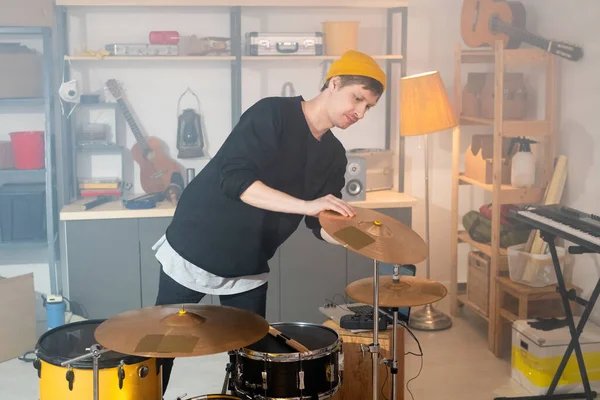 Contemporary young musician in casualwear bending over drum kit while checking one of cymbals before individual rehearsal in garage