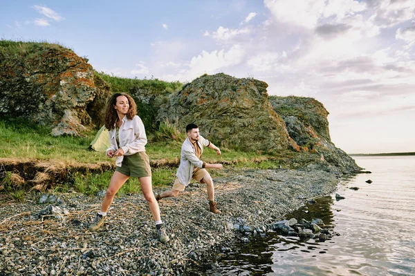 Happy young couple in boots, shorts and shirts throwing stones in water while standing on riverbank on background of rocks