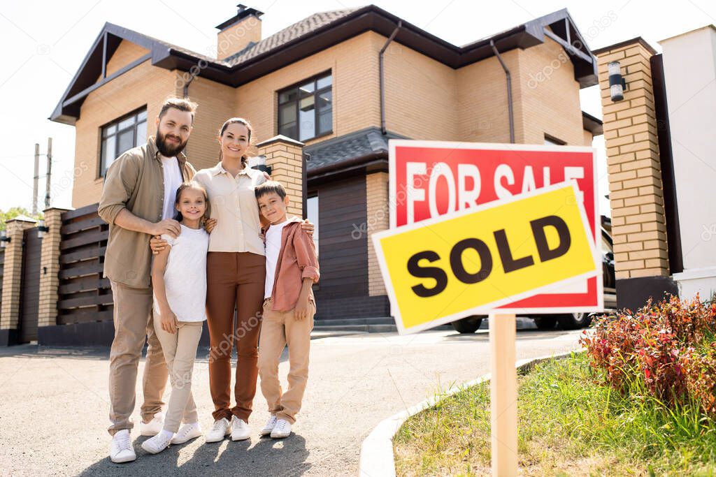 Portrait of smiling young family with two kids embracing each other against new house, moving into new house concept