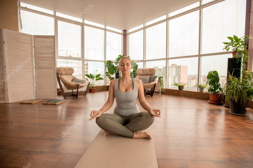 Young active blond sportswoman crossing legs while sitting on mat in pose of lotus and practicing meditation exercise in domestic room