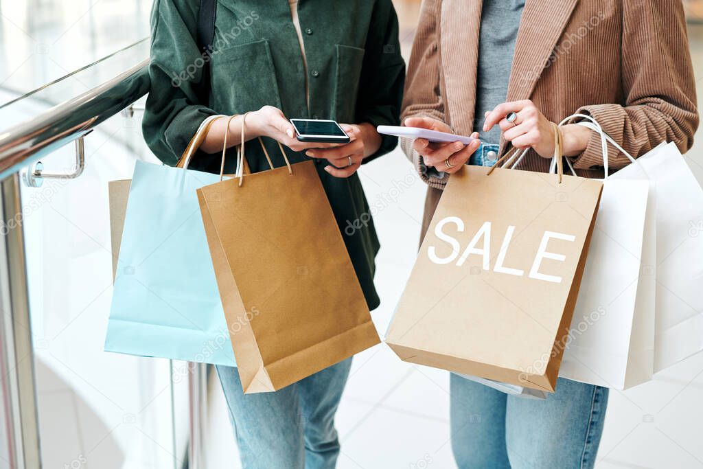 Hands of two young contemporary shoppers with smartphones and paperbags scrolling through online goods and comparing prices
