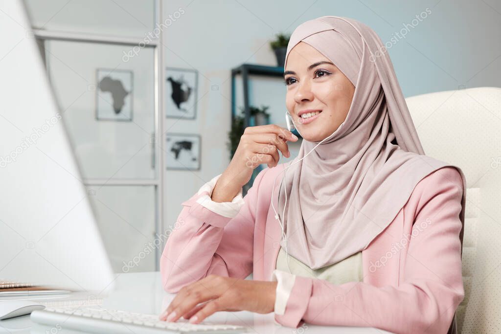 Portrait of dreamy pensive young Muslim woman sitting at cafe table, working on laptop and looking through window
