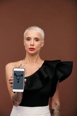 Kollarında ve göğsünde dövmeleri olan çağdaş zarif bir kadın ve ekranında transseksüel sembolü olan akıllı telefon taşıyan kısa sarı saçlı.