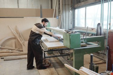 Genç ciddi fabrika işçisi, tezgahın üzerine eğildiği sırada ahşap tahtanın ya da atölyenin yüzeyini pürüzsüz hale getirmek için mekanik el aleti kullanıyor.