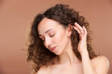 Genç, dingin bir kadın kameranın önünde dikilirken uzun, kahverengi saçlarına dokunuyor.