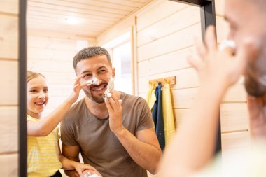 Genç, neşeli bir adam banyoda aynanın karşısında sevimli küçük kızıyla birlikte dururken sakalına köpük sürüyor.