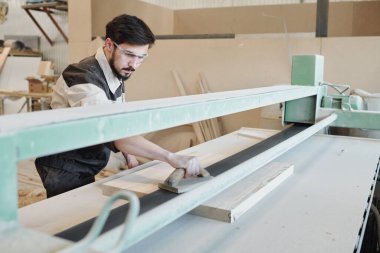 İş elbisesi içinde genç bir adam tezgahın üzerinde eğiliyor ve fabrikada çalışırken ahşap bir aletle çalışma yüzeyini düzleştiriyor.