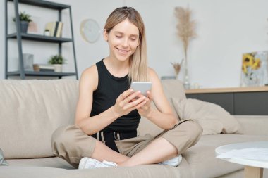 Rahat giyimli mutlu genç bir bayan bacaklarını kavuşturmuş bir şekilde kanepede oturuyor ve akıllı telefondan evde otururken fotoğraflara bakıyor.