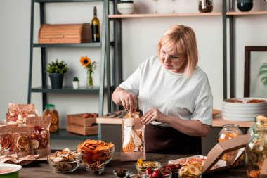 Olgun ev hanımı kurutulmuş portakal, armut, hurma, kivi ve çilekleri mutfak masasının yanında kağıt paketlere dolduruyor.