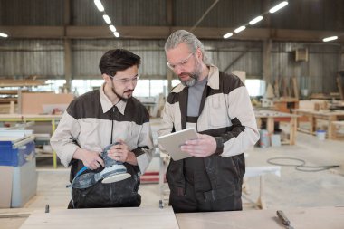 İş kıyafetleri ve koruyucu gözlük takan genç stajyer ahşap işçiliğin öğütücü yüzeyi yanında duruyor.