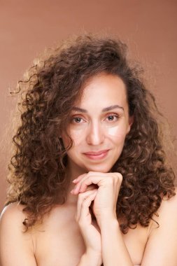 Göz altı bölgesiyle ilgilenen güzel, genç esmer kadın bej arka planda ellerini çenesinden ayırmıyor.