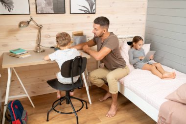 Günlük giysili genç adam tablet kullanarak okul kızıyla yatakta otururken tahta masa başında küçük oğluna ödevlerinde yardım ediyor.