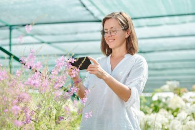 Gözlüklü ve beyaz gömlekli gülümseyen olgun bir kadın diğer bitkilerin arasında dikilirken akıllı telefon kamerasıyla çiçeklerin fotoğrafını çekiyor.