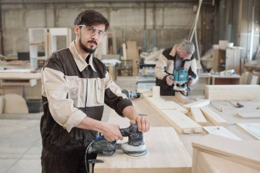 Genç fabrika işçisi öğütücü kullanarak işyeri yüzeyini pürüzsüz hale getiriyor ve daha fazla işlem için ahşap tahtayı hazırlıyor.