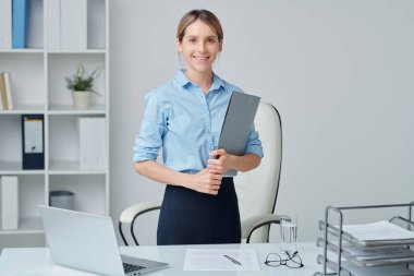 Resmi giyimde genç başarılı iş lideri elinde mali belge tutarken koltuğa ve raflara karşı ayakta duruyor.