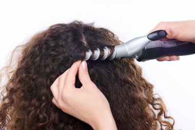 Genç esmer bayanın kafasının arkasında uzun siyah saçları, beyaz arka planda kuaför tarafından elektrikli aletle kıvrılmış.