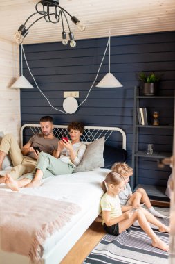 Anne babalar yatakta dinlenirken ve çocukları yatak odasında yerde otururken çağdaş aile mobil cihazlarla kayıyor.
