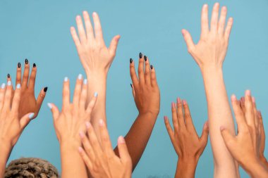 Çeşitli etnik kökenlere sahip, beyaz ve koyu tenli geniş bir grup genç kadın, mavi arka plana karşı kameranın önünde kollarını kaldırıyor.