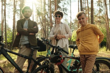 Üç kişilik aile kameranın önünde kırsal alanda çam ağaçlarının gövdesine karşı bisiklet sürüyor ve hafta sonları dinleniyor.