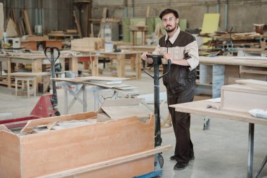 Modern fabrikanın genç işçisi atölye boyunca ilerlerken büyük bir kutu dolusu atölye artıklarıyla el arabası itiyor.