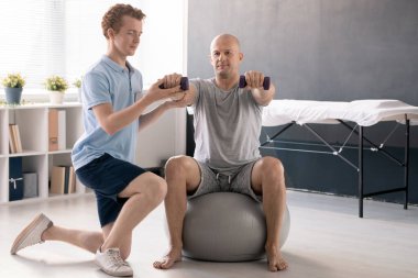 Spor kıyafetleri içindeki genç fizyoterapist erkek hastaya halterlerle egzersiz yapması ve spor yaparken sağ kolunu desteklemesi için yardım ediyor.