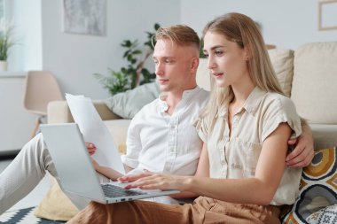 Genç, ciddi sarışın koca, televizyon izlerken karısının yanındaki koltukta oturmuş dizüstü bilgisayarına bakıyor.