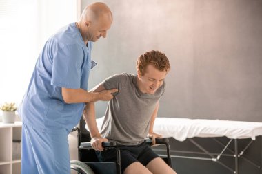 Üniformalı kel klinisyen, rehabilitasyondan sonra kolunu ve dirseğini desteklerken genç bir adamın tekerlekli sandalyeye oturmasına yardım ediyor.