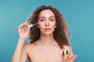 Koyu uzun dalgalı saçlı genç bir kadın elinde küçük bir kavanoz ve pipetle yüzüne gençleştirici serum sürüyor.