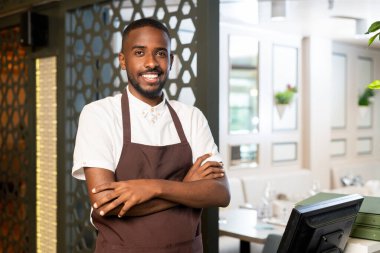 Afrika kökenli üniformalı neşeli bir garson dişlek bir gülümsemeyle ve restoran ortamında kollarını kavuşturarak sana bakıyor.