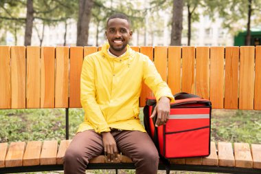 Sarı ceketli neşeli erkek kurye parkta bankta otururken müşterilerin siparişleriyle birlikte büyük kırmızı sırt çantasını tutuyor.