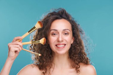 Yüzünde üç kozmetik fırça tutan dişlek bir kadın mavi arka plana profesyonel makyaj uygulayacak.