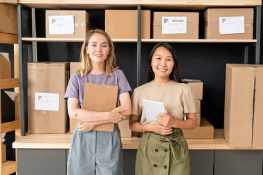 Çevrimiçi dükkanların depolama odasındaki iki neşeli genç işçi, ellerinde paketlenmiş kutularla büyük ahşap rafların yanında duran belgelerle.