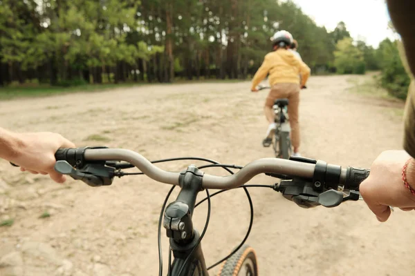 緑の木々に囲まれた田舎道を進む息子を追いながら自転車のハンドルを握る若者の手 — ストック写真