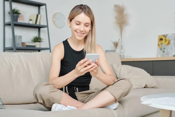 幸せな若いです女性でキャミソールに座って彼女の足が交差し 家にいながらスマートフォンで写真を見て — ストック写真