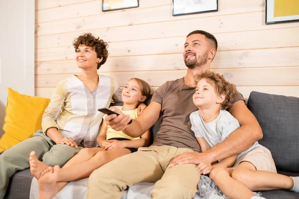 Genç, neşeli bir çift ve günlük giysiler içinde iki küçük çocuk oturma odasında kanepede oturup hafta sonları televizyon izliyorlar.