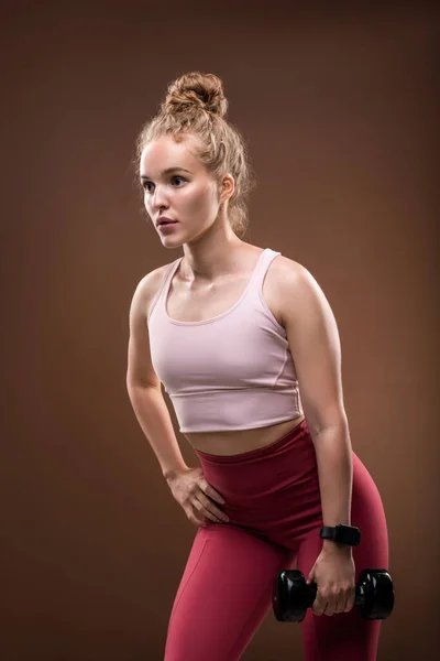アクティブなブロンド女の子でスポーツウェア保持ダンベルで左手でザ右1オンヒップ上の物理的な運動中に茶色の背景 — ストック写真